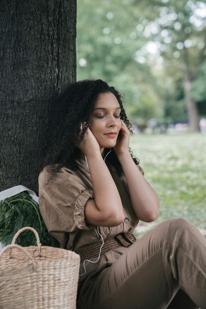 A woman leaning against a tree in a park, listening to music, oblivious to wild coincidences.