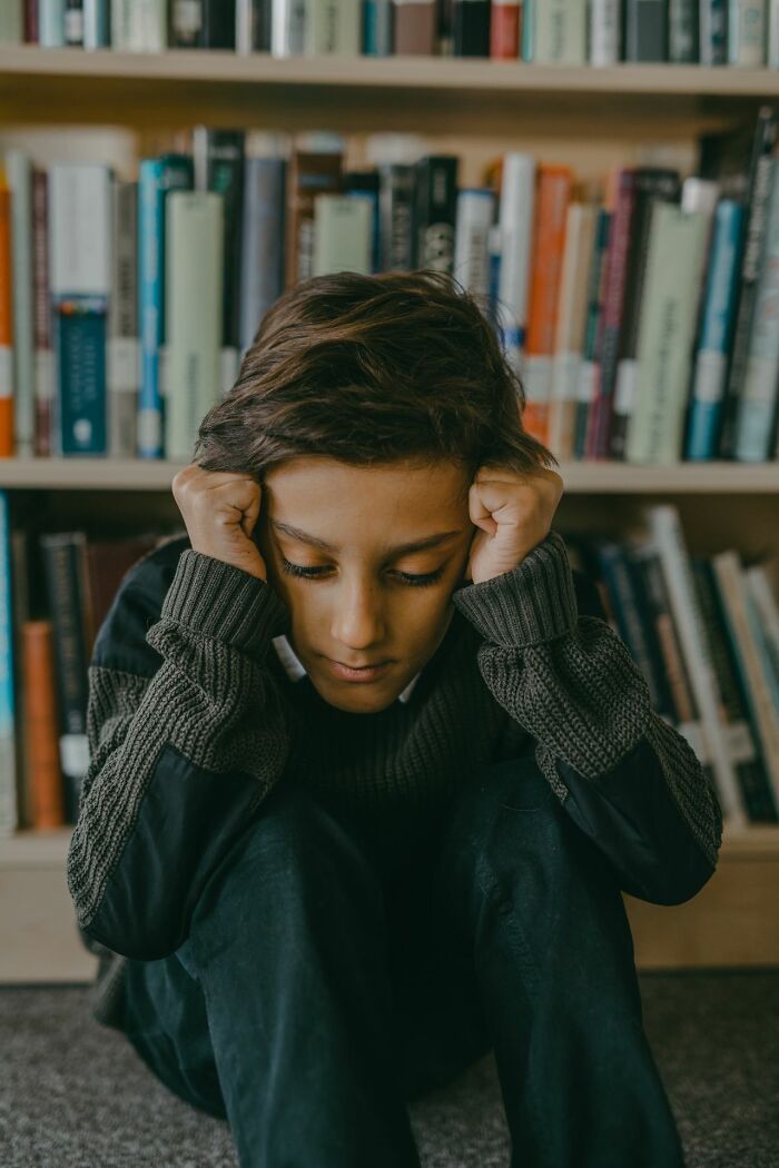 A young boy sits on the floor, head in hands, in front of a bookshelf, reflecting emotions in polygamist families.