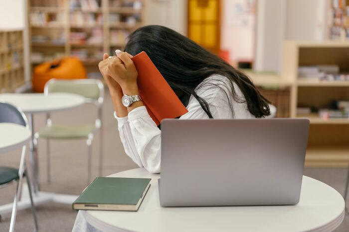 A young woman in an awkward situation, hiding her face with a book in a library.