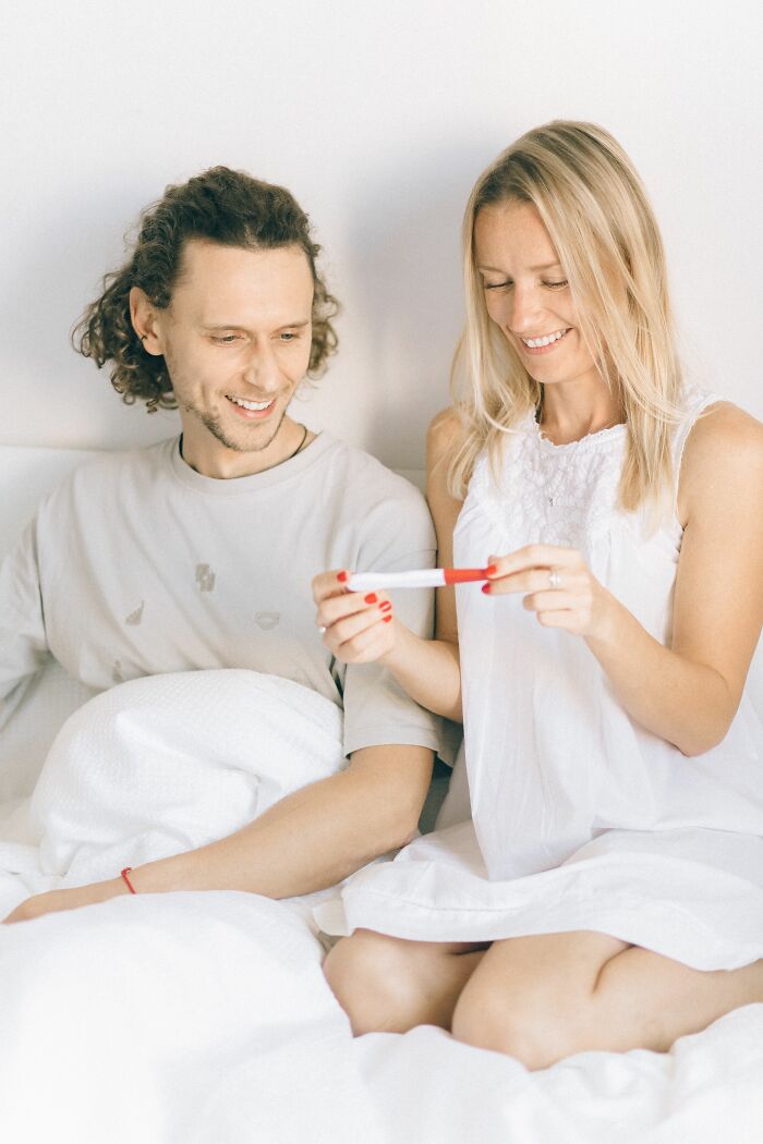 A couple smiling on a bed, looking at a pregnancy test. Learning NSFW facts about human bodies.