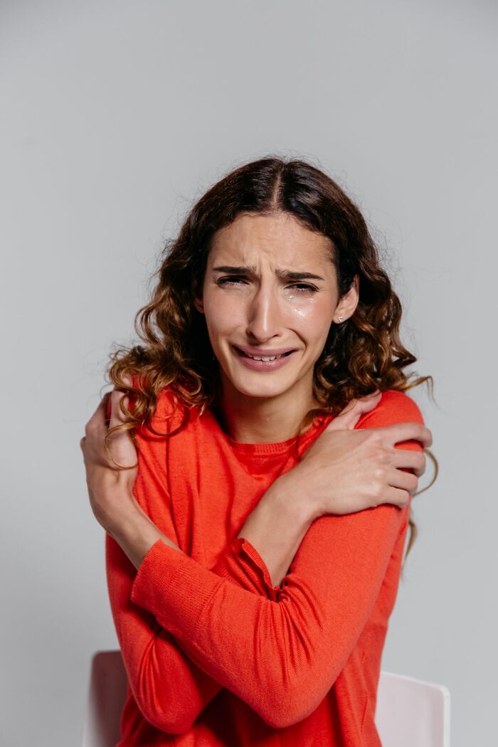 A woman with curly hair in a red sweater cries, hugging herself, depicting deep emotional pain.