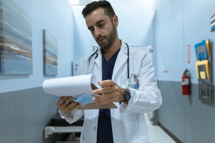 A doctor in a white coat looking at a clipboard in a hospital hallway, potentially in an awkward situation.