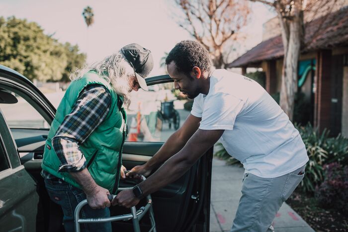 A man helps an elderly partner with a walker get into a car, showing a supportive moment to marry.