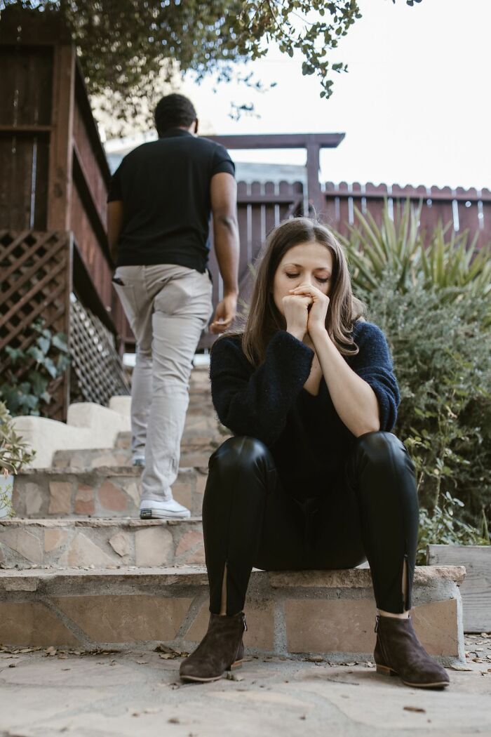 A woman sits on steps, head in hands, experiencing profound pain as a man walks away.