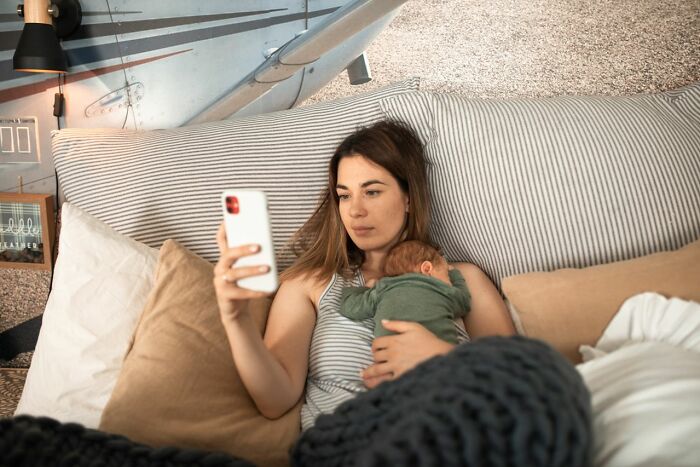 A mother holding a baby while looking at her partners phone in bed, indicating a discovery on the phone.