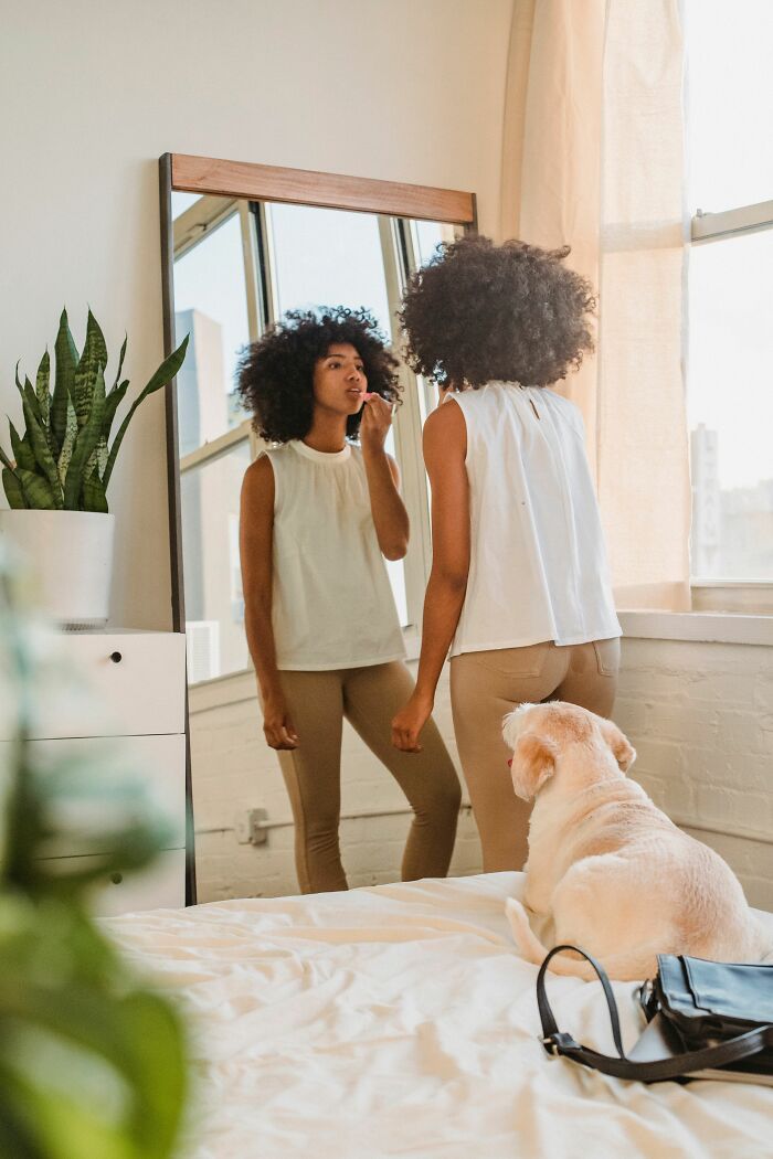 A woman applies lipstick in a mirror while her dog watches, representing human habits that confuse dogs.