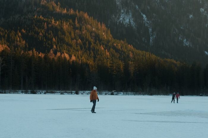 Person in orange jacket walking on icy snowfield near forested mountains