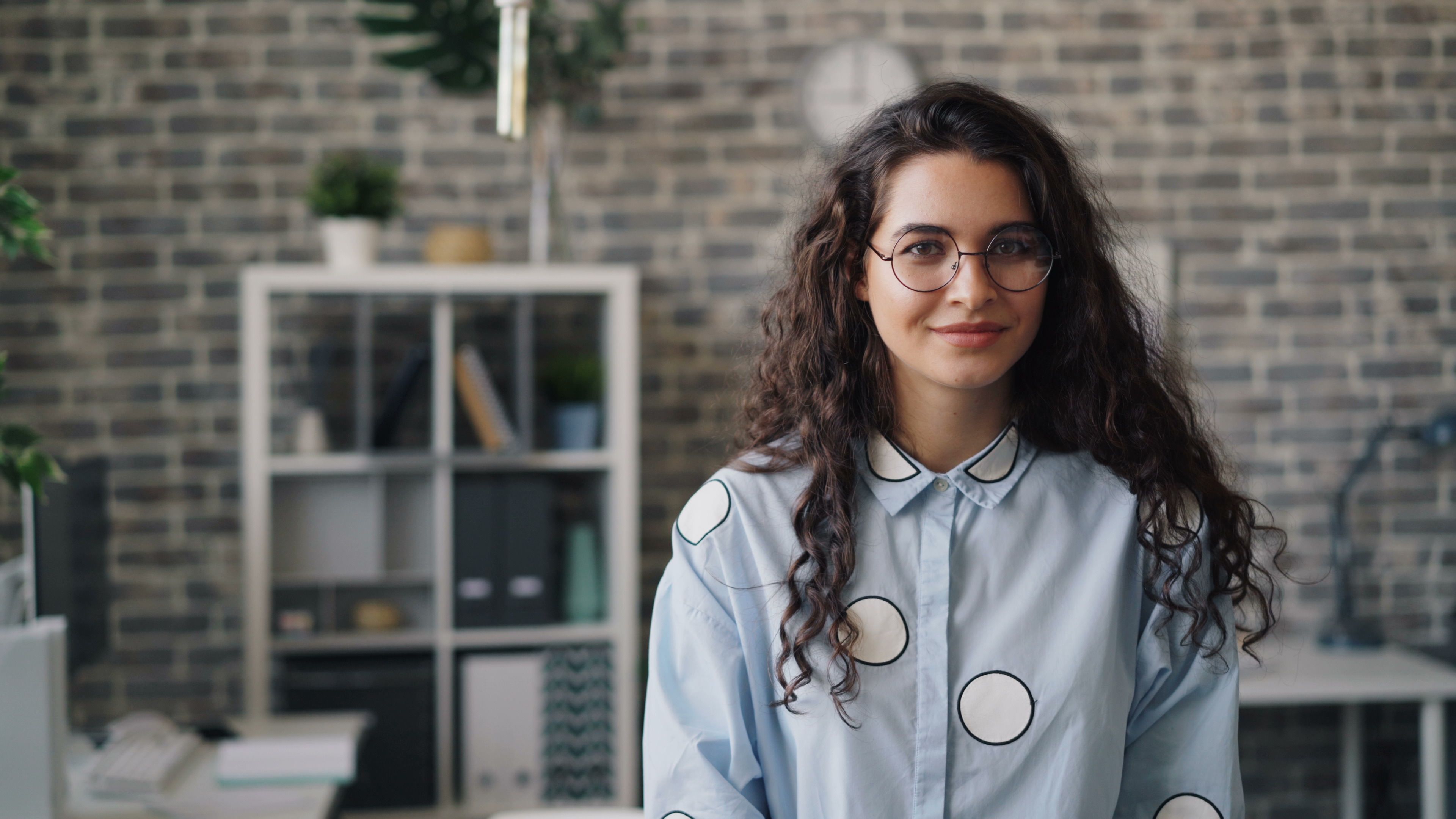 A woman with curly hair and glasses, breaking up with her ex, smiling slightly in an office setting.