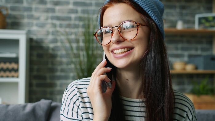 A young woman in glasses and a beanie smiling on the phone, showing men's turn offs women roll their eyes at.