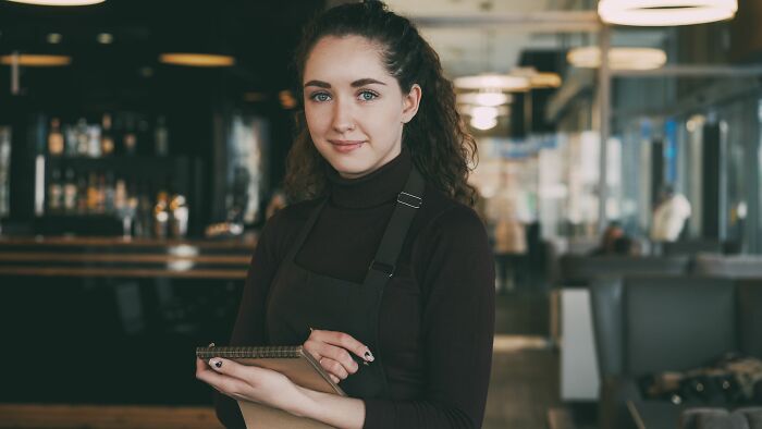 A young waitress in an apron holding a notepad, smiling, attentive to social cues.