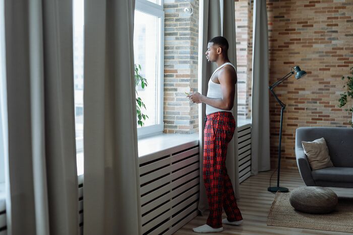 Man in pajamas standing by window drinking tea indoors