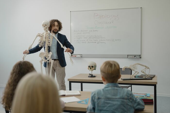 Biology teacher explaining human skeleton to students in classroom with skeleton model Biology teacher explaining human skeleton to students in classroom with skeleton model