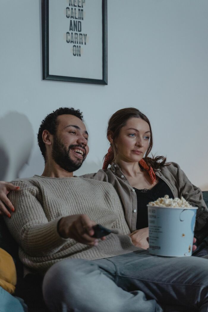 A couple watching TV, the man laughing and the woman looking displeased, possibly due to things found on her partners phone.
