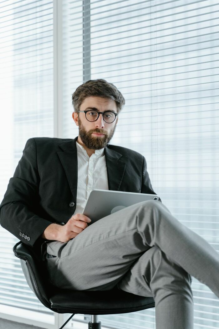A man in glasses and a suit holds a tablet, showing hints of being very smart, sitting by a window.