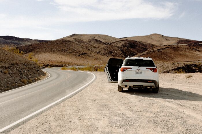 A white SUV with its door open on a dirt road next to a winding paved road, with barren hills in the background. A wild coincidence.