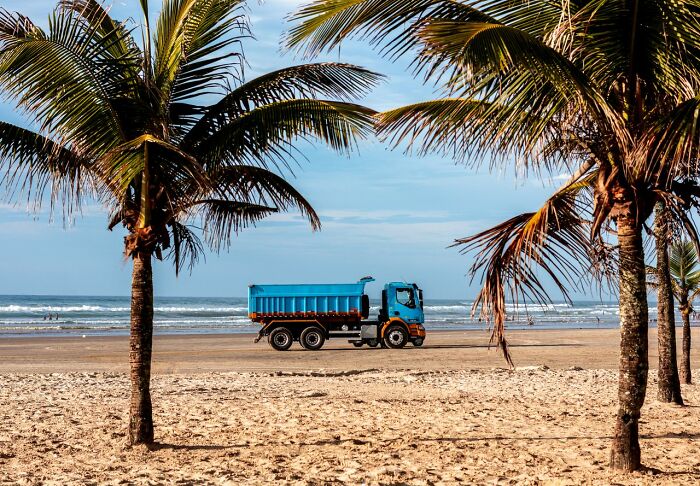 A blue dump truck driving on a sandy beach with palm trees, illustrating a crazy fact about Wyoming's two escalators.