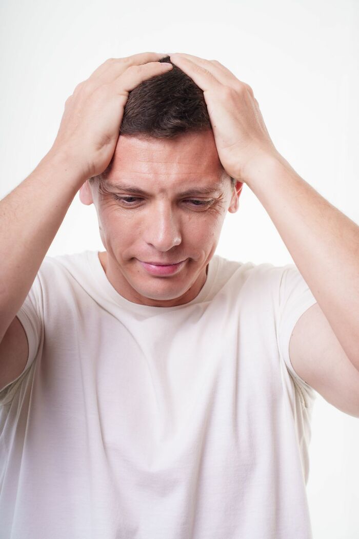 A man in a white t-shirt holding his head with both hands, looking down with a concerned expression, symbolizing an unserious ER visit.