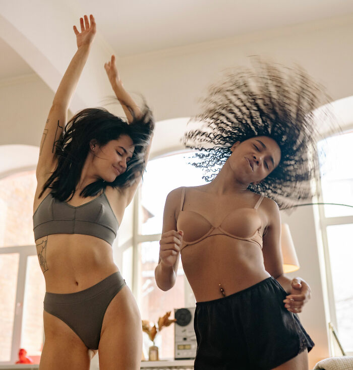 Two young women dancing joyfully at home, showcasing a fashion trend.