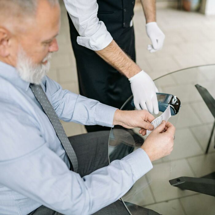 Man reading receipt while waiter in gloves serves, depicting entitled parents and service