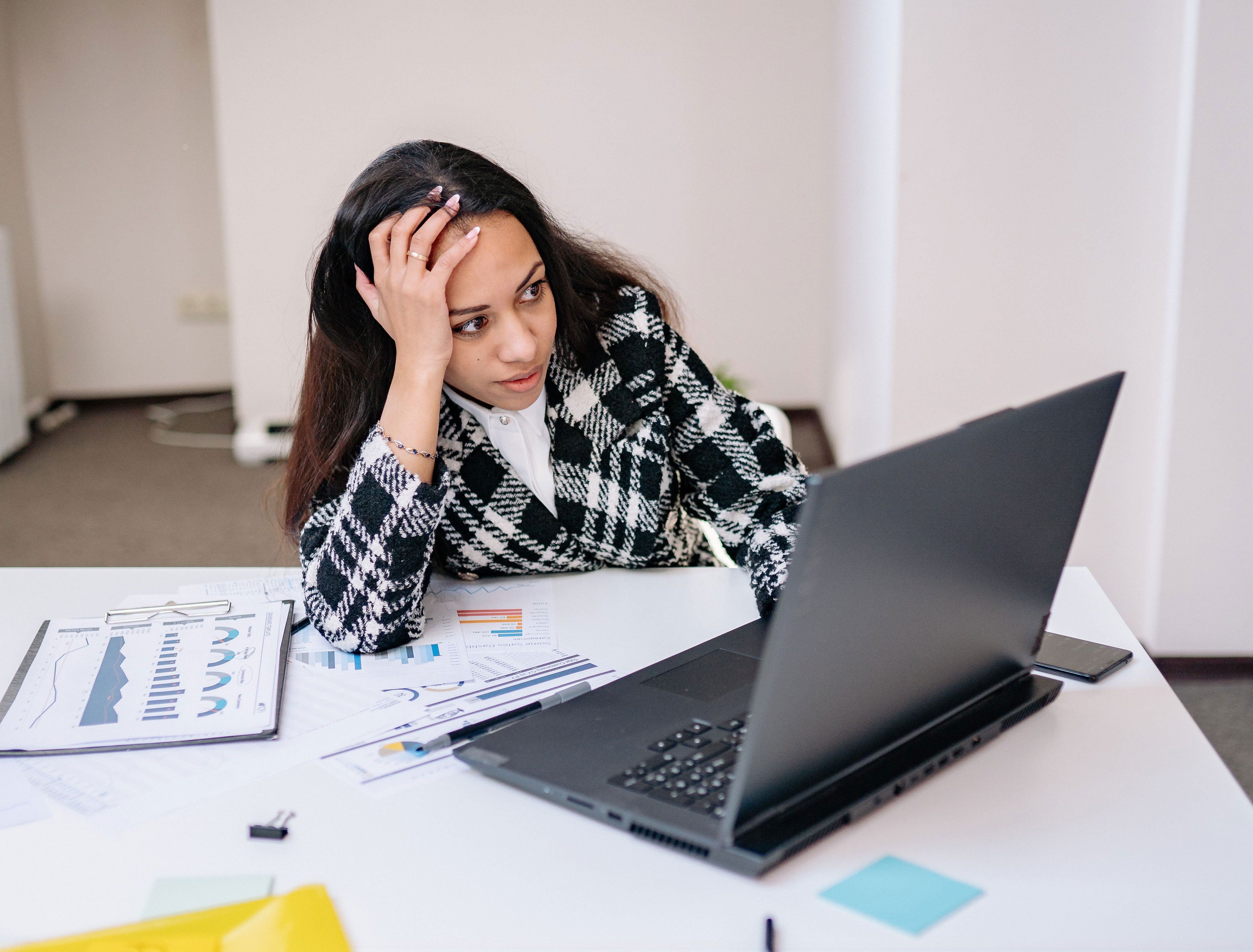 A frustrated woman in a plaid jacket sits at a desk looking at a laptop, demonstrating the stress from an ableist Karen.