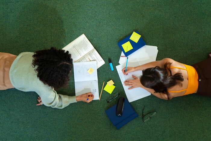 Two students lying on a green floor, studying together for general knowledge questions to beat an above-average IQ.