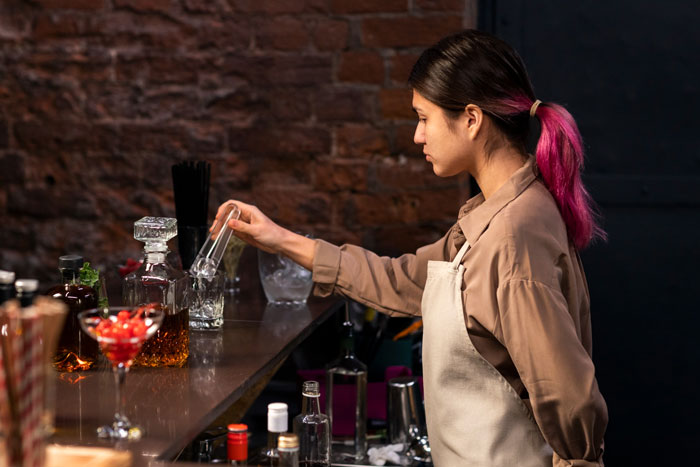 A pregnant woman with pink hair working in a pub, mixing a drink behind the bar.