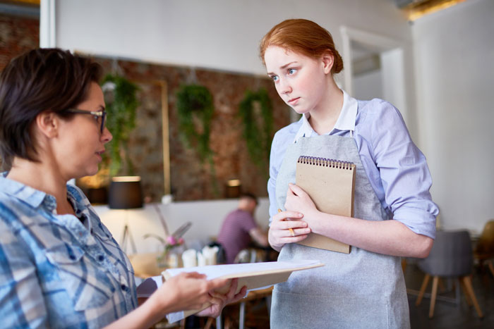 A pregnant woman, a pub worker in an apron, listens with a confused expression to an older woman. Religious fanatics fume.