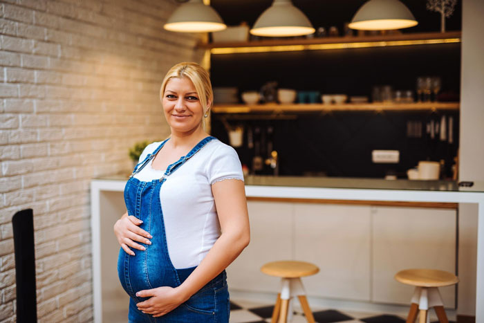 A pregnant woman, wearing denim overalls and a white t-shirt, smiles while working in a pub.