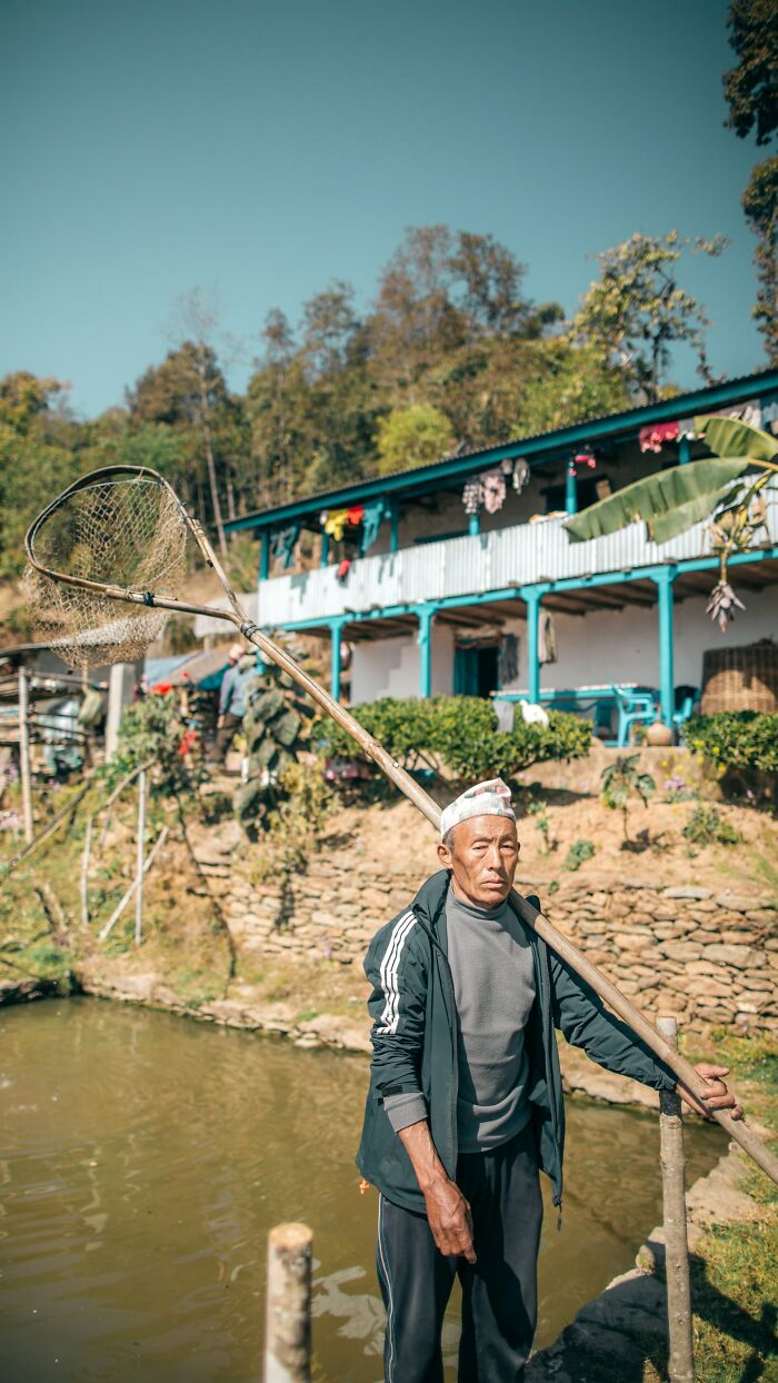 An older man with a fishing net stands by a pond near a house, representing stories from polygamist families.