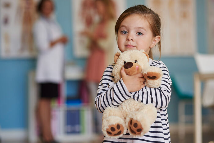 Primary girl hugging a teddy bear in a doctor's office with blurred adults