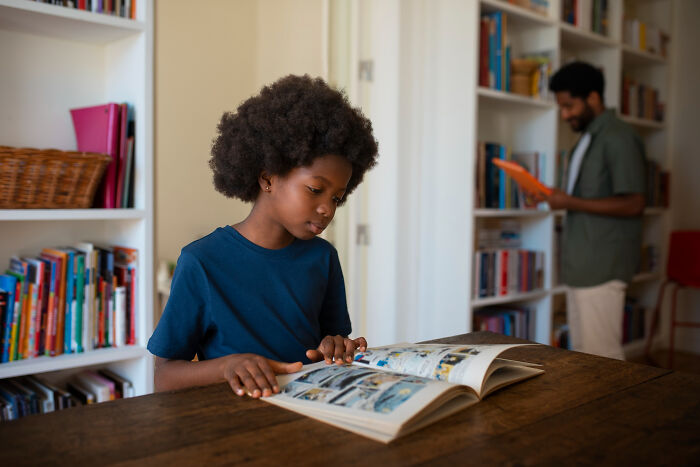 Primary kid reading a book in a library with an adult in the background