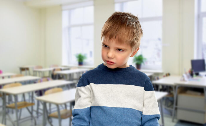 Angry primary school boy standing in classroom with empty desks and windows