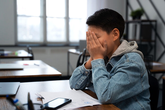 Distressed primary school boy covering face with hands sitting at desk