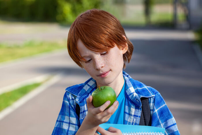 Red-haired primary kid holding green apple looking thoughtfully