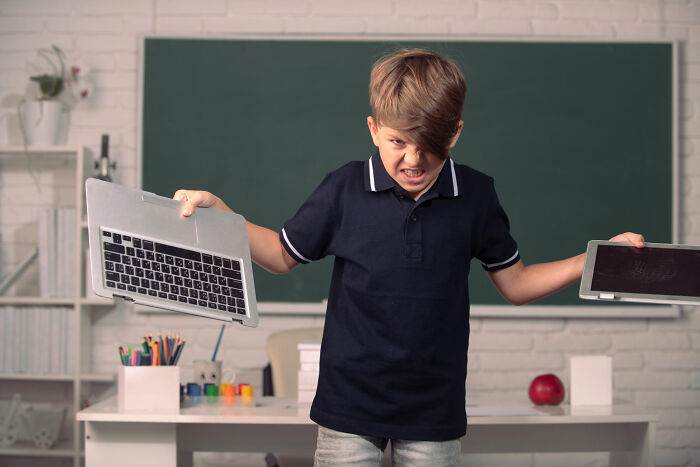 Frustrated primary boy angrily holding broken laptop parts in a classroom