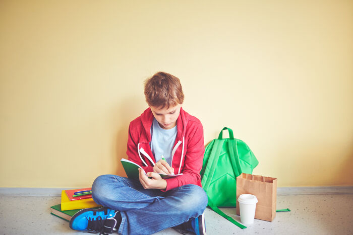 Primary school boy writing in notebook sitting on floor with backpack and lunch bag