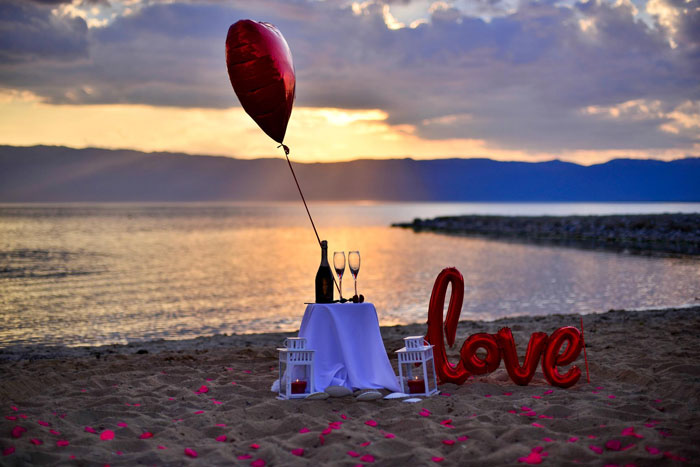 Romantic beach proposal setup with fake petals, champagne, and love balloons at sunset.