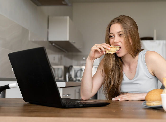 Woman eating snack while watching laptop in kitchen