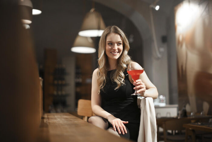 Woman in black dress holding drink at bar with confident smile