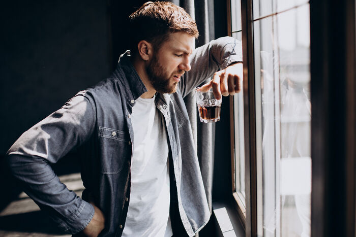 Man looking troubled holding drink by window in dim room
