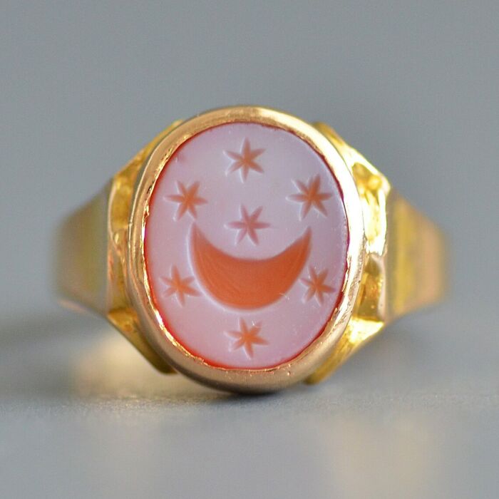 Intact after years, a gold ring featuring a pinkish-white oval stone engraved with a crescent moon and stars.