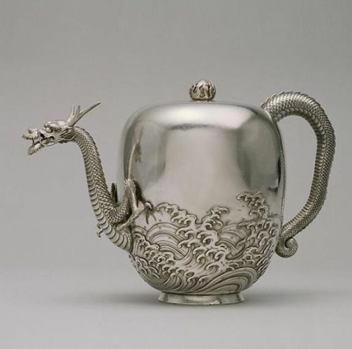 An elaborate silver teapot with a dragon handle and spout, an intact object from many years ago, featuring intricate wave patterns.