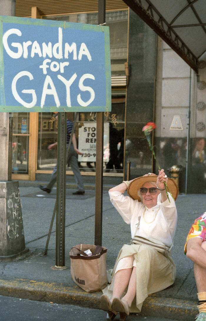 An older woman with a straw hat sitting by a sign that says Grandma for Gays, an interesting object still intact.