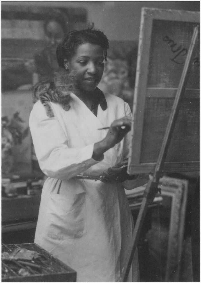 A black and white photo of a woman in an apron painting on an easel, an interesting object still intact after all these years.