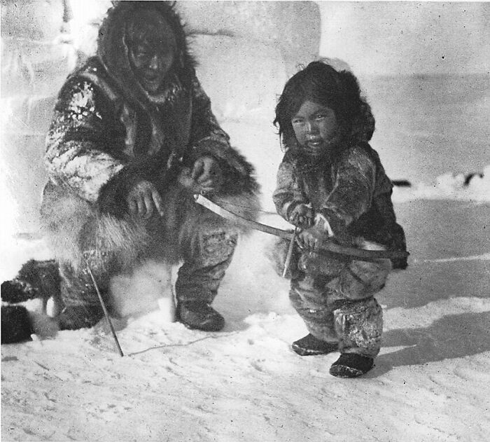 An Inuit adult and child in warm clothing, holding a bow and arrow in the snow, a rare historical photo.