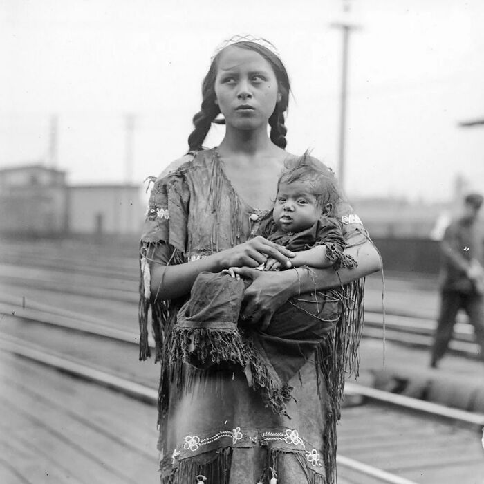 A young Indigenous woman holding a baby, standing near railroad tracks, in a historical photo.