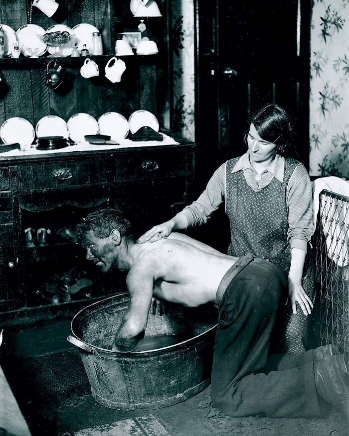 A woman giving a man a bath in a tub in a historical photo of a simple home interior.