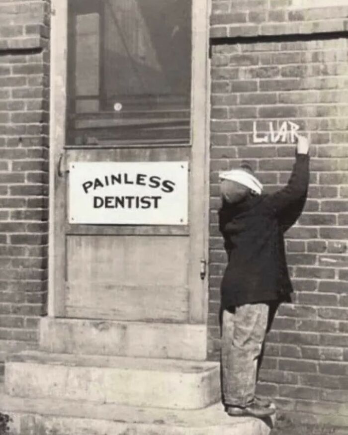 A black and white historical photo of a child writing 'LIAR' on a brick wall next to a 'PAINLESS DENTIST' sign.