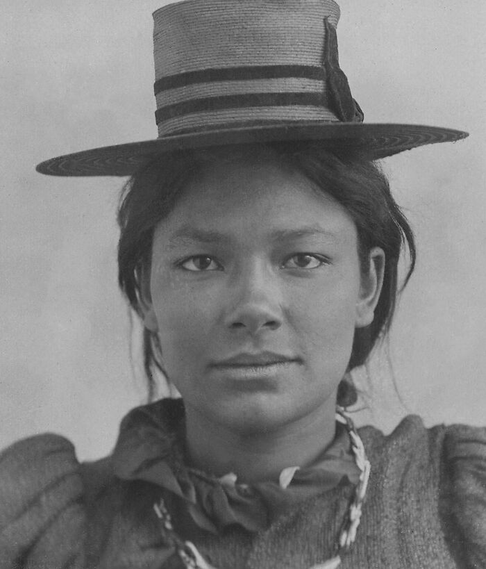 A black and white historical photo of a young indigenous woman wearing a wide-brimmed hat and a beaded necklace.