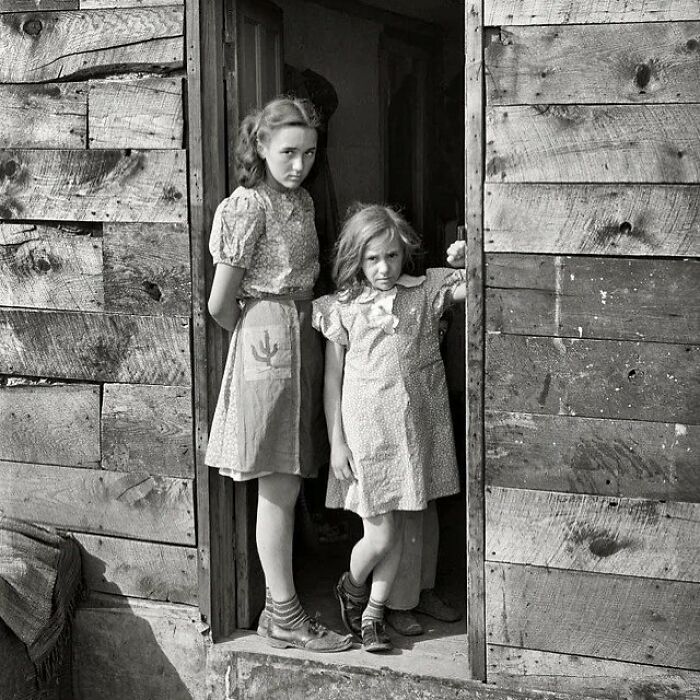 Two young girls in plain dresses standing in a doorway of a rustic wooden building, a rare historical photo.
